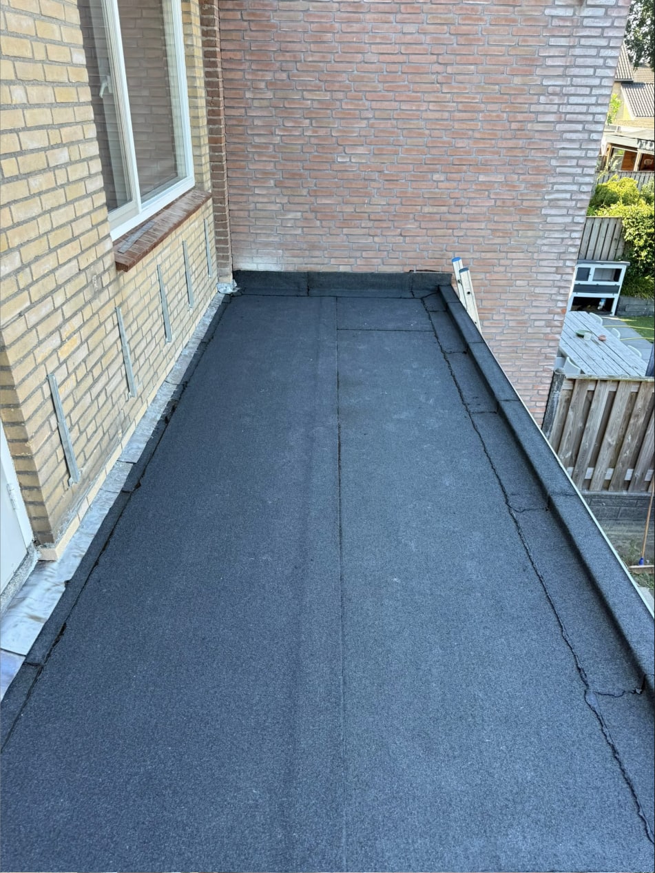 Afgewerkt plat dak met nieuwe bitumen toplaag langs een bakstenen gevel